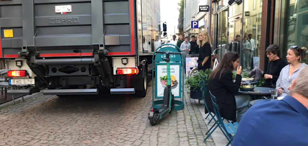 Bred lastbil på smal gata med folkliv